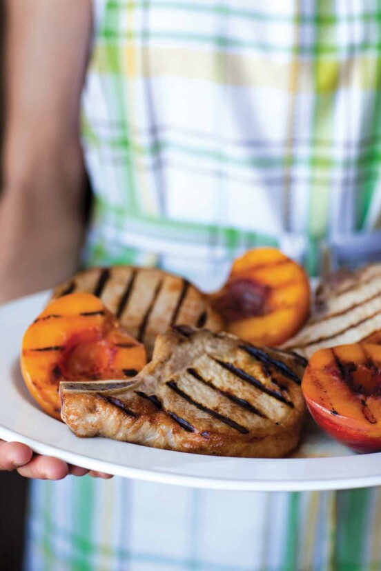 A woman holding a white platter of grilled country pork chops with grilled peaches.