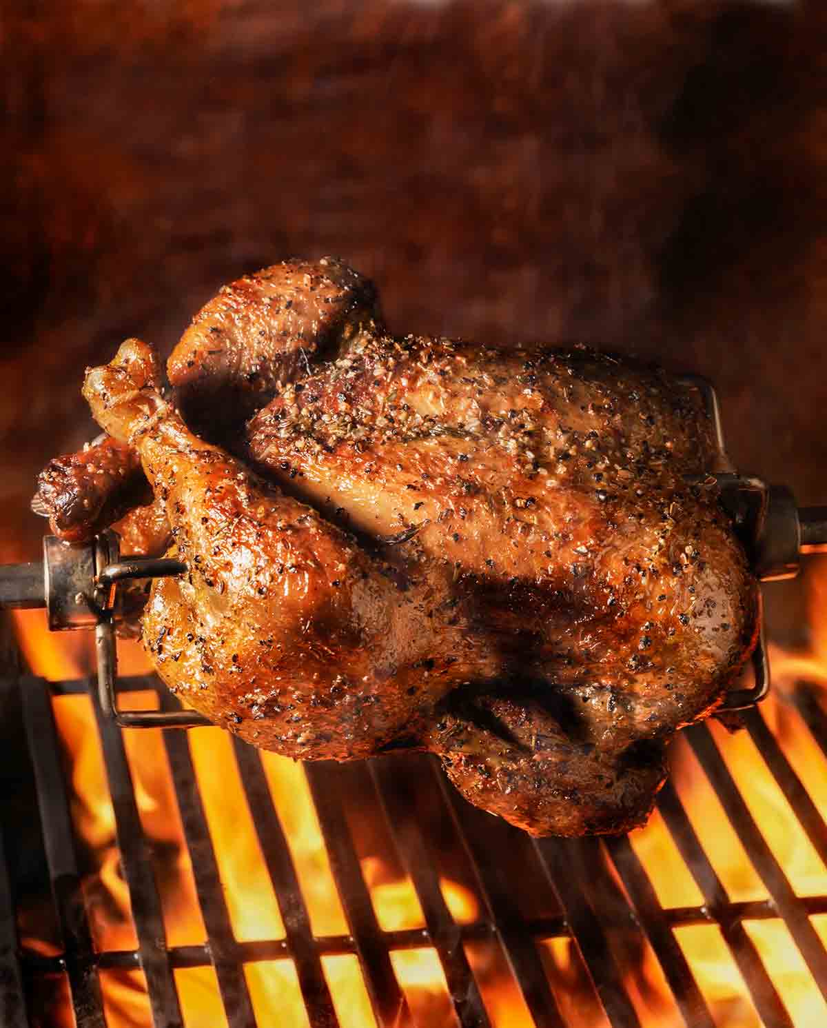 A rotisserie chicken suspended over a grill with flames below.
