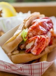 A lobster roll in a cardboard sleeve with a little lettuce and a lemon wedge in the background.