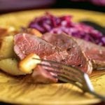 Slices of cider-basted venison with roasted potatoes and red cabbage on a yellow plate.