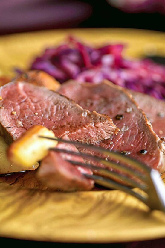 Slices of cider-basted venison with roasted potatoes and red cabbage on a yellow plate.