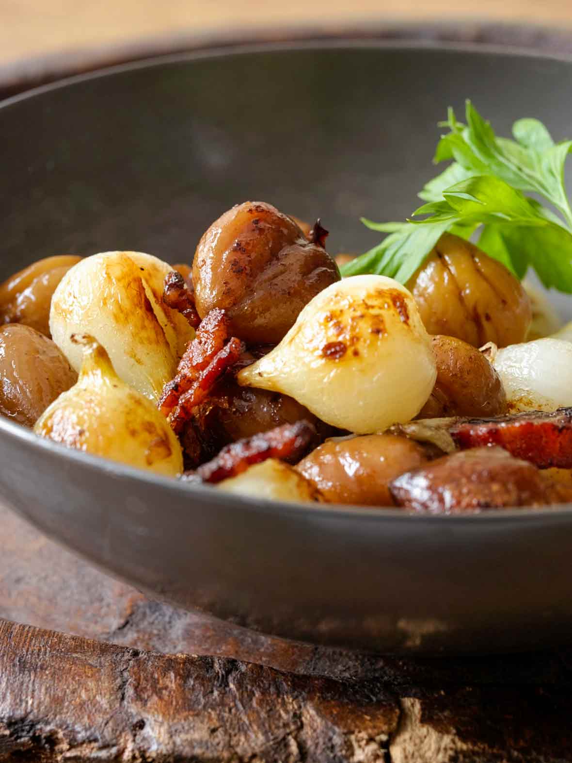 A grey ceramic bowl filled with Portuguese onions, chestnuts, and bacon, topped with an Italian parsley garnish.