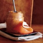 A jar of roasted applesauce with a spoon resting inside on a saucer with a strip of apple peel.