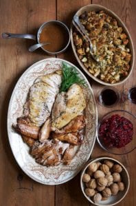An oval platter with roasted and braised turkey, a dish of stuffing, a bowl of cranberry sauce, a bowl of chestnuts, two glasses of wine, and a small pot of gravy on a wooden table.