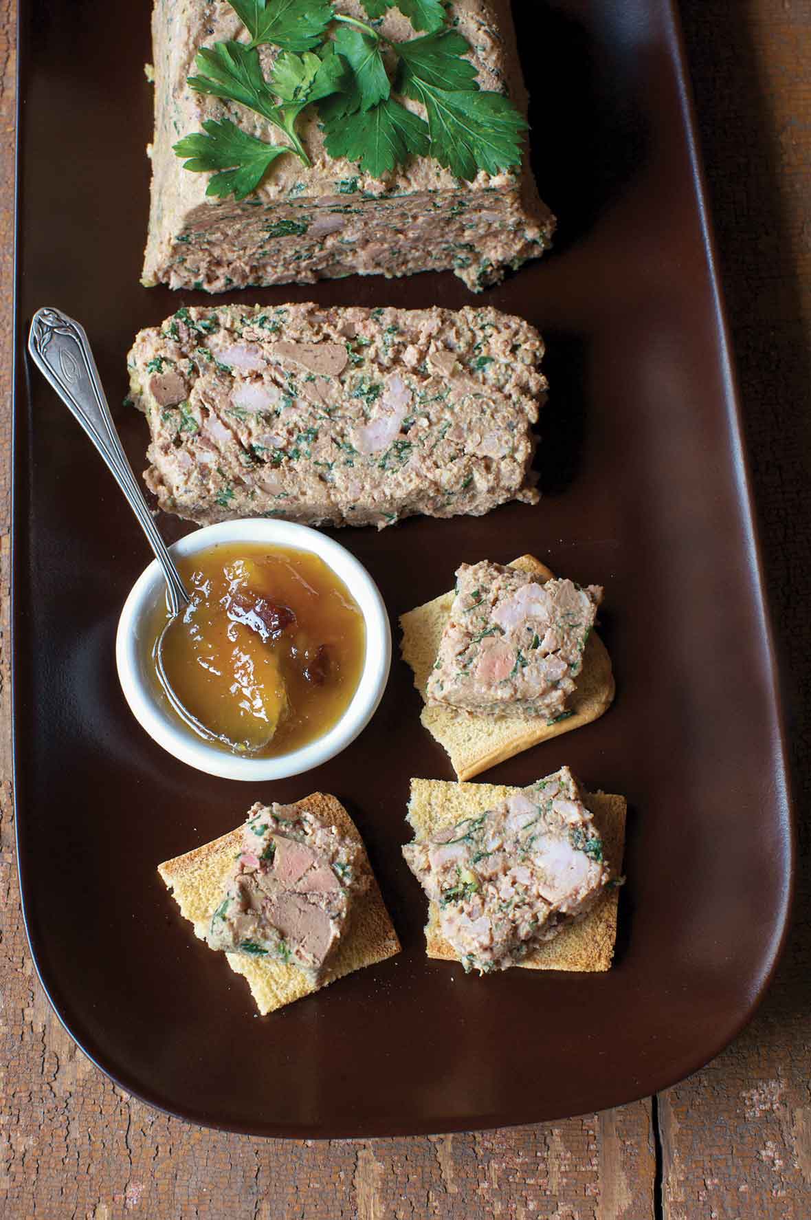 Chicken liver pate on a wooden tray with slices on toast beside a bowl of chutney.