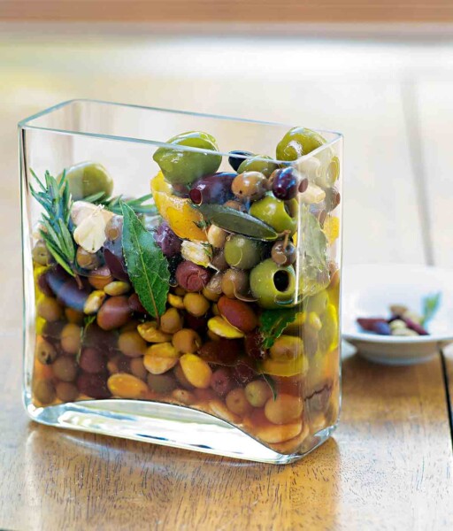 A square glass container with herb-marinated olives, bay leaves, rosemary, and thyme.