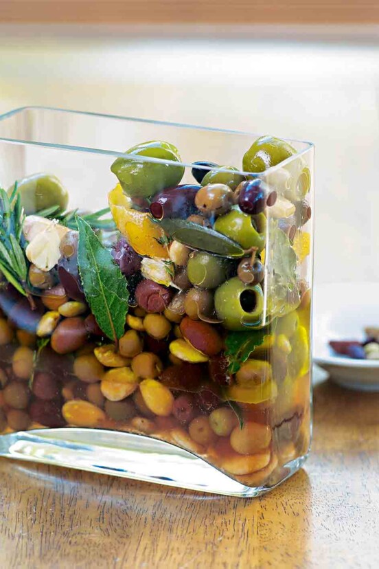 A square glass container with herb-marinated olives, bay leaves, rosemary, and thyme.