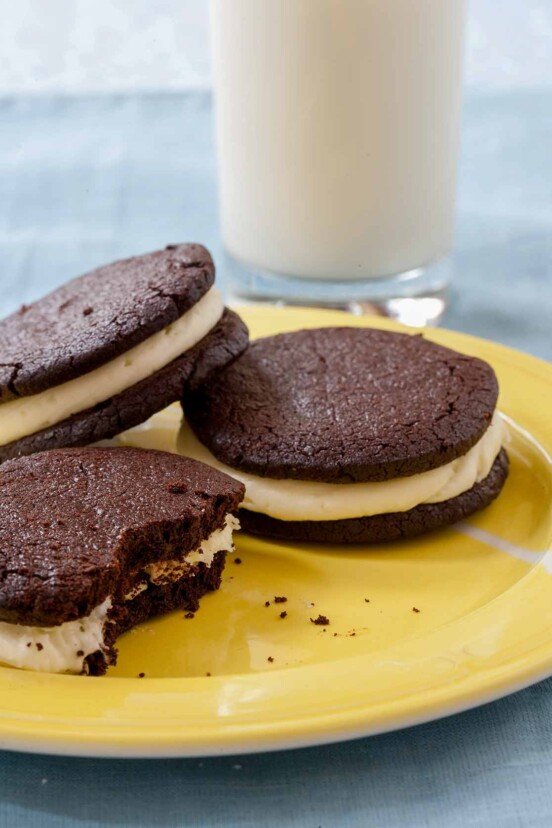 Homemade Oreos piled on a yellow plate beside a glass of milk.