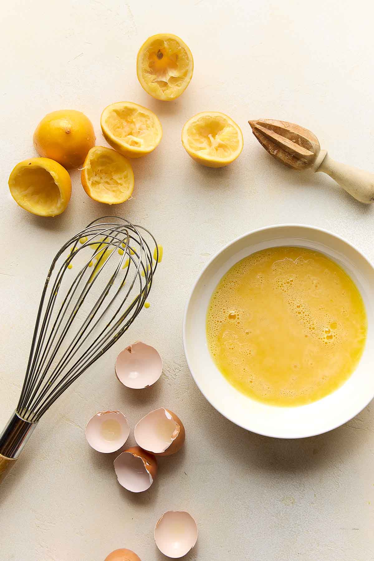 A bowl of uncooked Meyer lemon curd, egg shells, and juiced lemon halves, and a whisk.