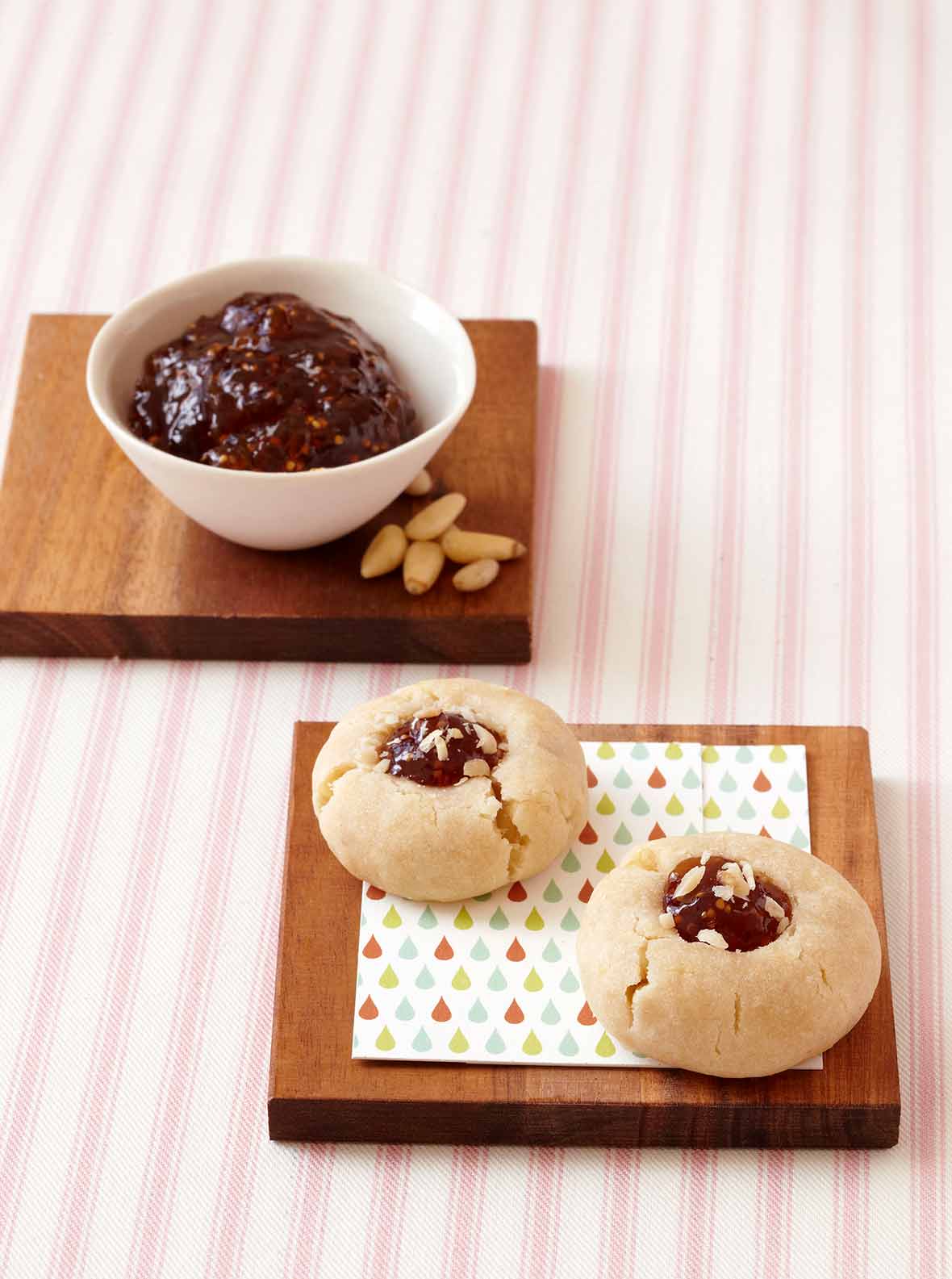 Mix-and-match thumbprint cookies on a wooden trivet in front of a bowl of jam and a few pine nuts.