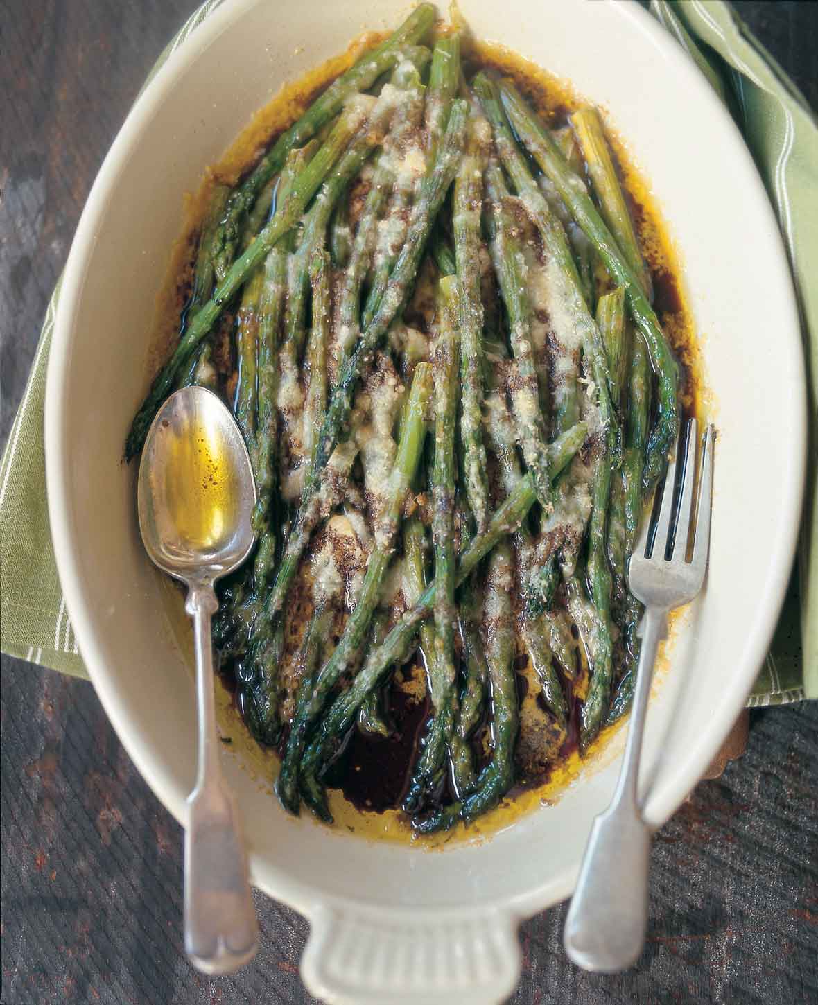 An oval serving dish filled with baked asparagus with Parmesan and balsamic vinegar with a fork and spoon resting on top.