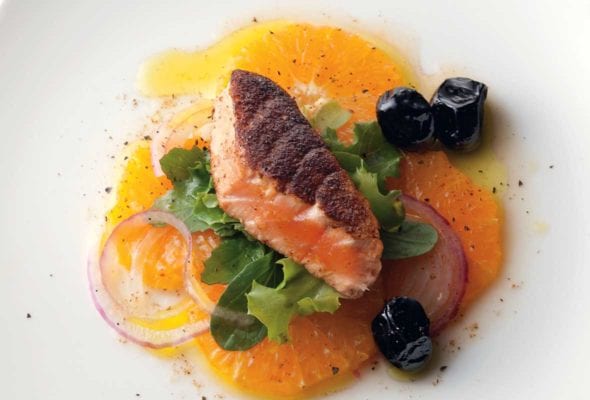 A white plate with a piece of pan-seared Moroccan salmon on a bed of orange slices, red onion, and greens with three black olives on the side.