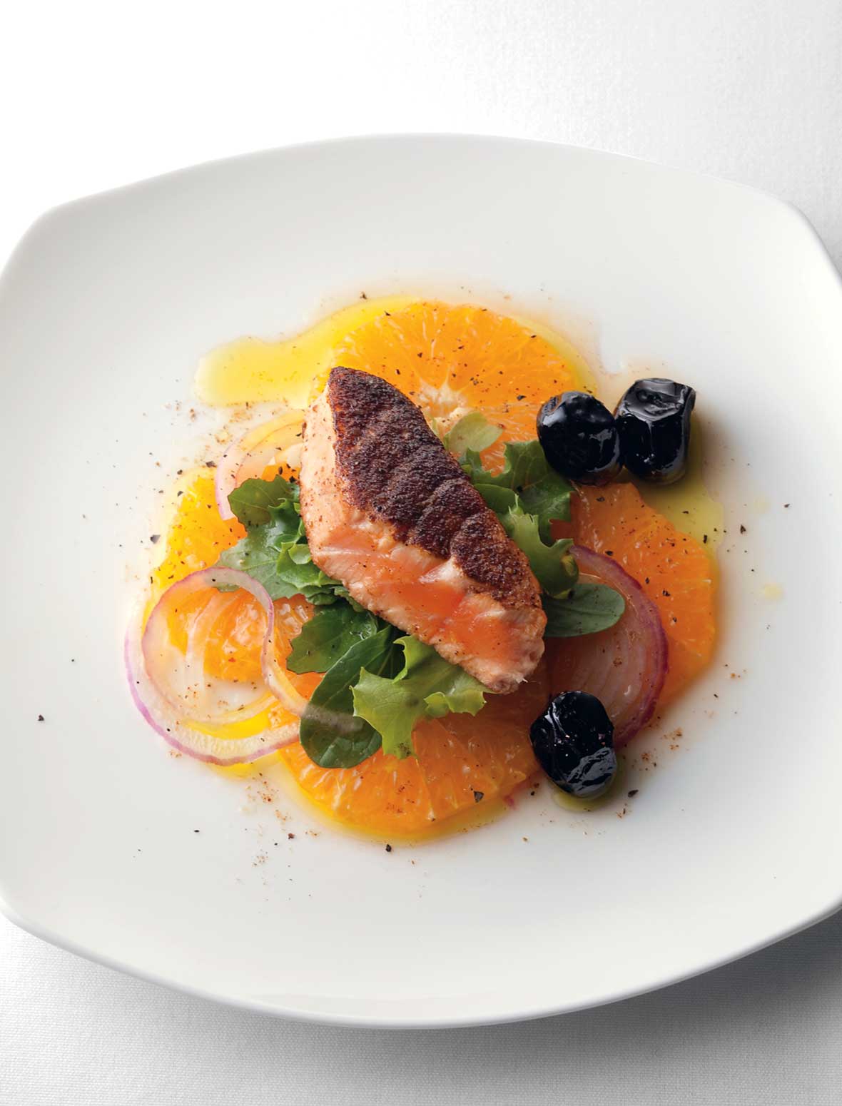 A white plate with a piece of pan-seared Moroccan salmon on a bed of orange slices, red onion, and greens with three black olives on the side.