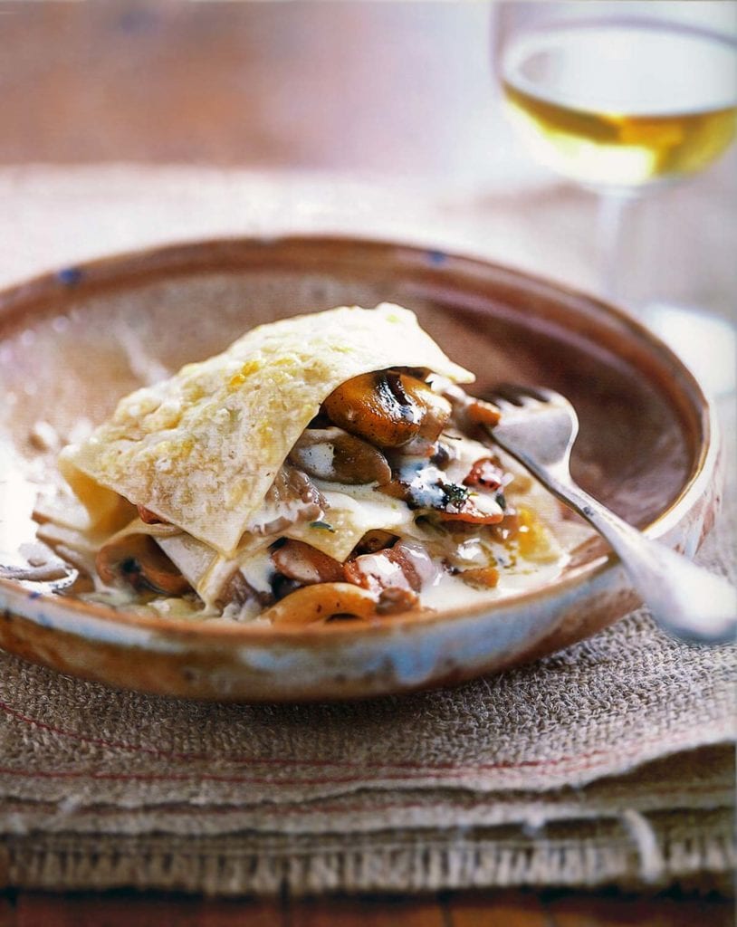 An earthenware bowl with an open lasagna of mushrooms, pine nuts, and thyme in it and a fork resting in the bowl and a glass of white wine in the background.