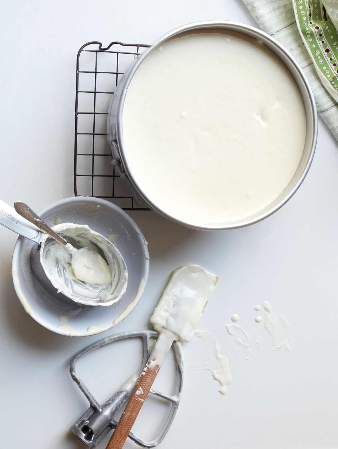 A New York–style cheesecake in a springform pan on a wire rack with a dirty bowl, spoon, spatula, and paddle attachment lying beside it.