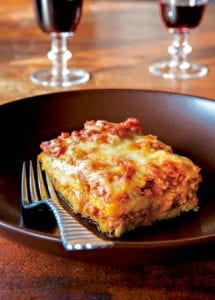 A square of polenta with layers of meat sauce on a plate with a fork, two glasses of wine