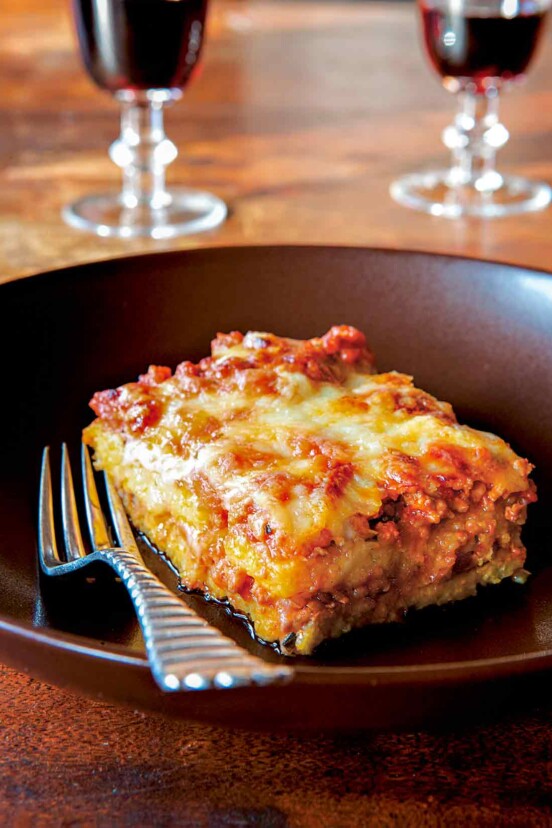 A square of polenta with layers of meat sauce on a plate with a fork, two glasses of wine