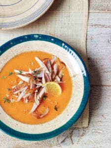 A blue-rimmed porcelain bowl filled with carrot soup topped with chicken and thyme and a lemon wheel.