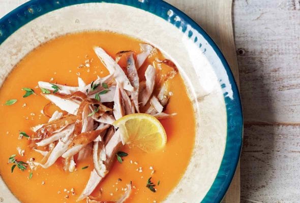 A blue-rimmed porcelain bowl filled with carrot soup topped with chicken and thyme and a lemon wheel.