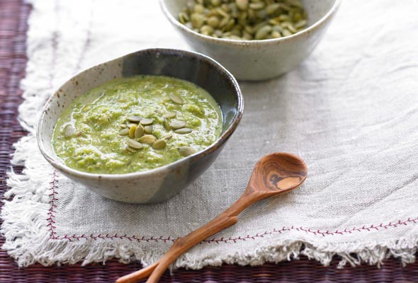 Pumpkin seed mole in a small ceramic bowl with a rustic wooden spoon, sitting on a linen tablecloth with a bowl of pepitas in the background.