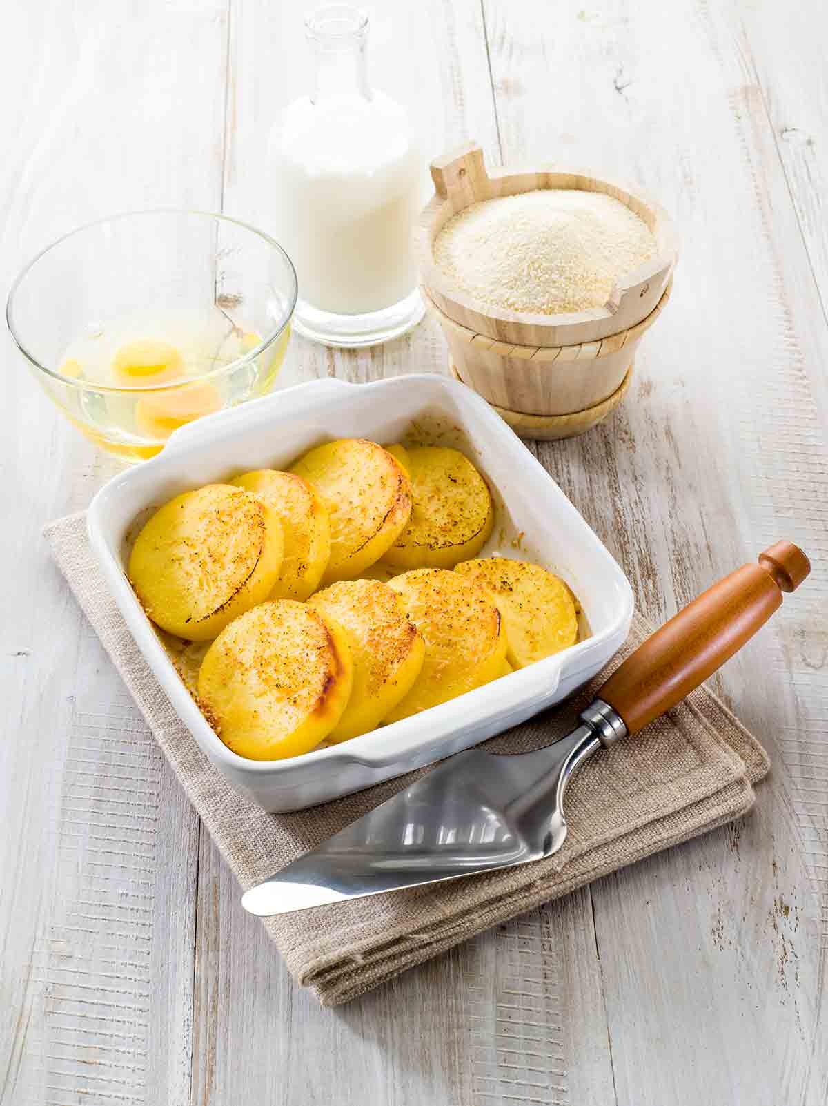 A white casserole dish filled with slices of semolina gnocchi or gnocchi alla Romana on a stack of linen napkins with a serving utensil beside it.