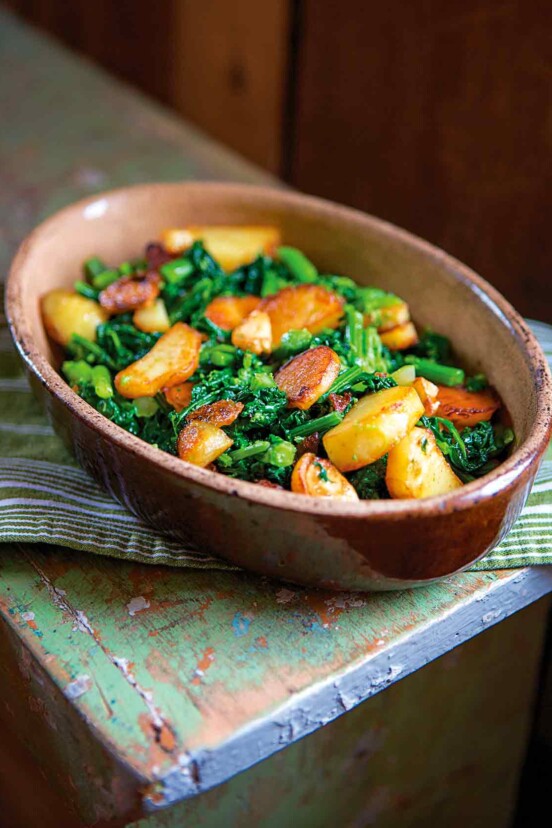 Pieces of sauteed broccoli rabe and slices of seared potatoes in a brown oval dish on a green bench.