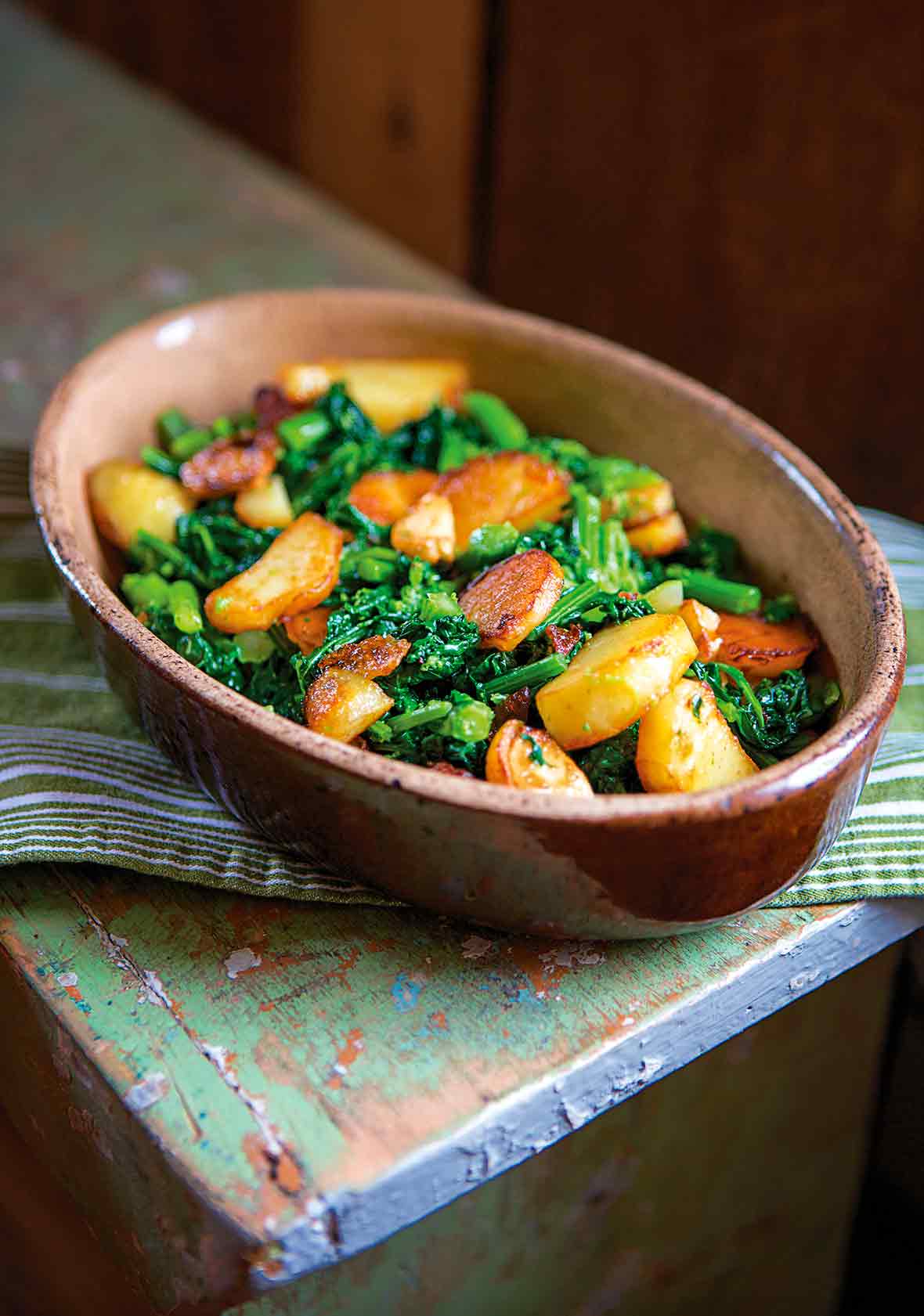 Pieces of sauteed broccoli rabe and slices of seared potatoes in a brown oval dish on a green bench.