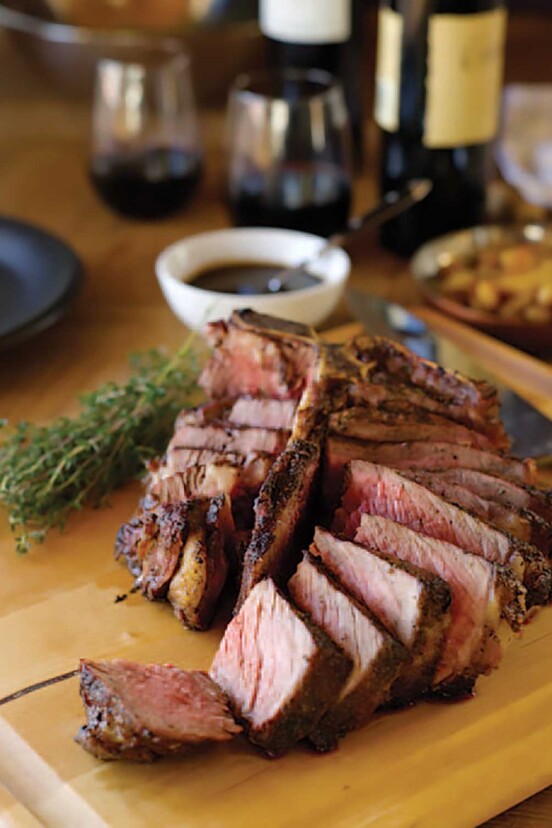A grilled porterhouse steak sliced on a wooden cutting board, flanked by fresh thyme and a white bowl of steak sauce, a bottle of red wine, and two glasses of wine.