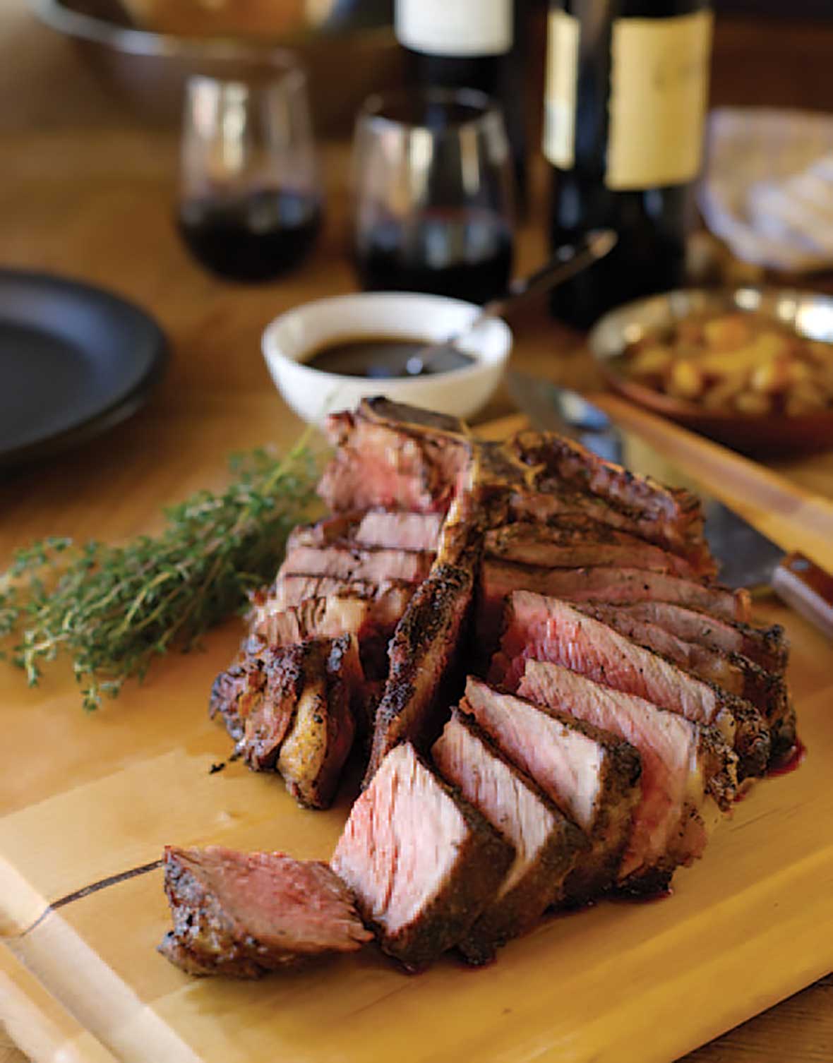 A grilled porterhouse steak sliced on a wooden cutting board, flanked by fresh thyme and a white bowl of steak sauce, a bottle of red wine, and two glasses of wine.