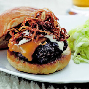 A Michael Schlow burger topped with cheese and caramelized onions on a white plate with a serving of slaw on the side.