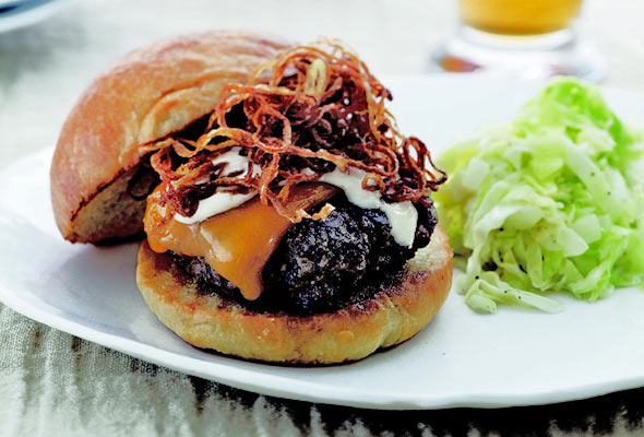 A Michael Schlow burger topped with cheese and caramelized onions on a white plate with a serving of slaw on the side.