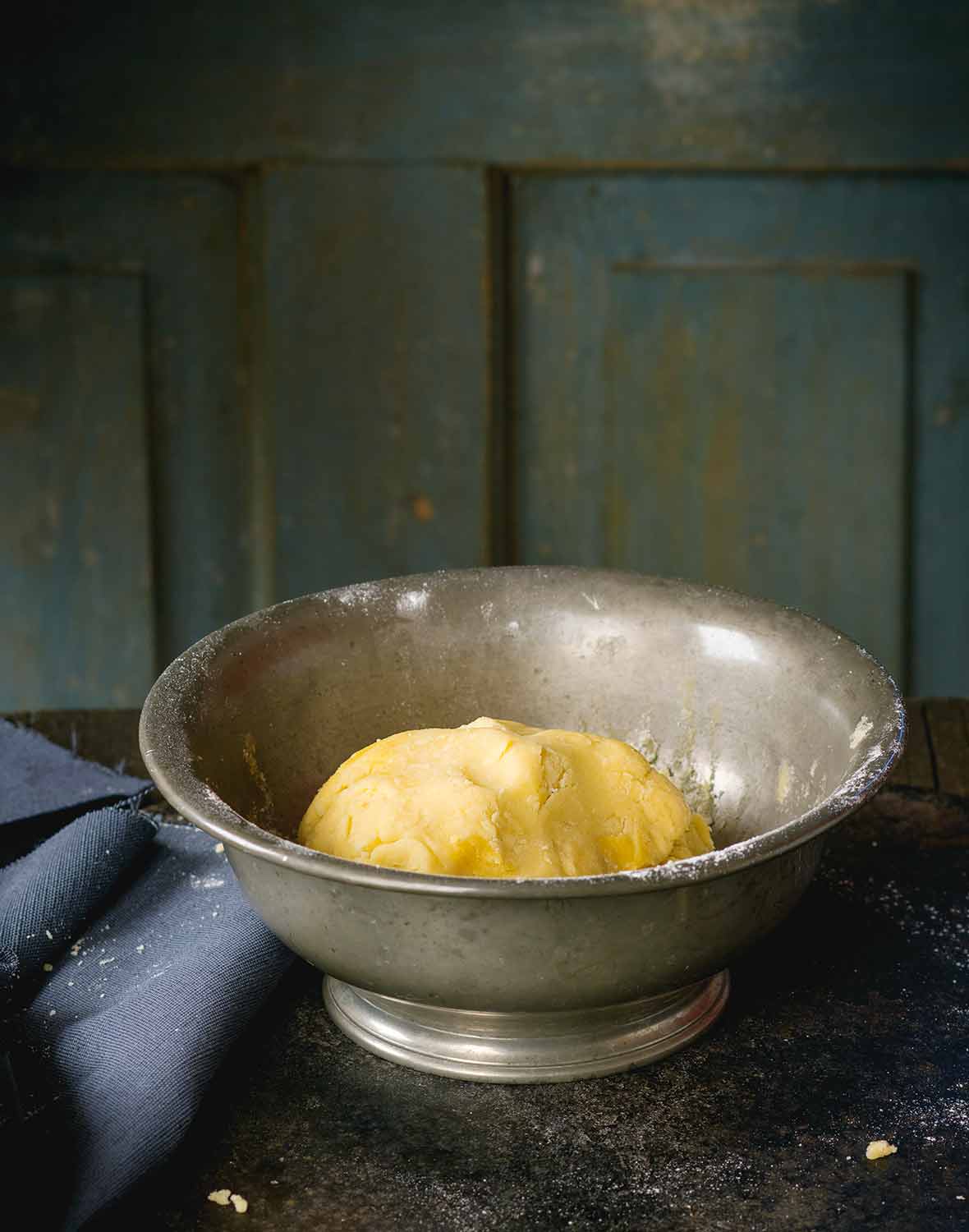 A ball of pâte sucrée in a metal bowl on a wooden table with a napkin next to it.