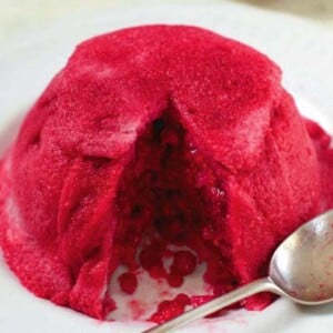 A bright pink summer pudding with one piece missing, sitting on a white plate with a spoon.