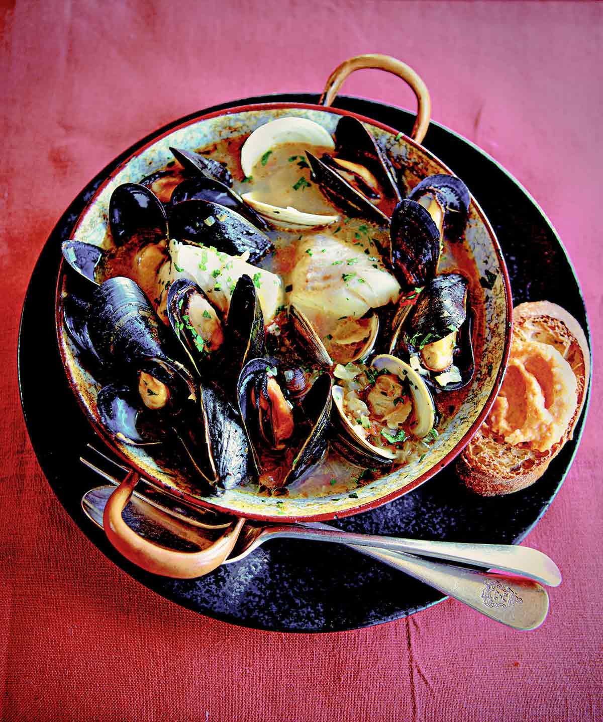 A dish filled with New England bouillabaisse for one with a fork and spoon resting beside it on a black plate.