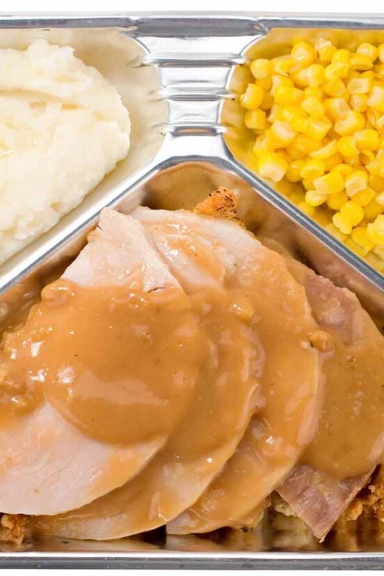 A tv dinner with sliced meat, stuffing, gravy, corn, and mashed potatoes in a foil container.
