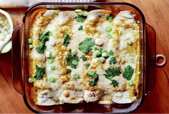 A glass baking dish with four chicken enchiladas topped with cheese, sliced scallions, and cilantro leaves.