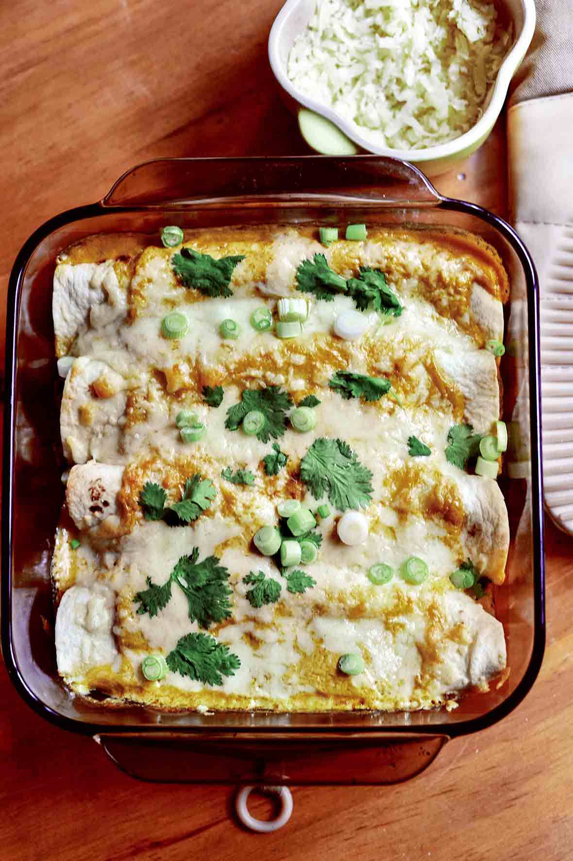 A glass baking dish with four chicken enchiladas topped with cheese, sliced scallions, and cilantro leaves.