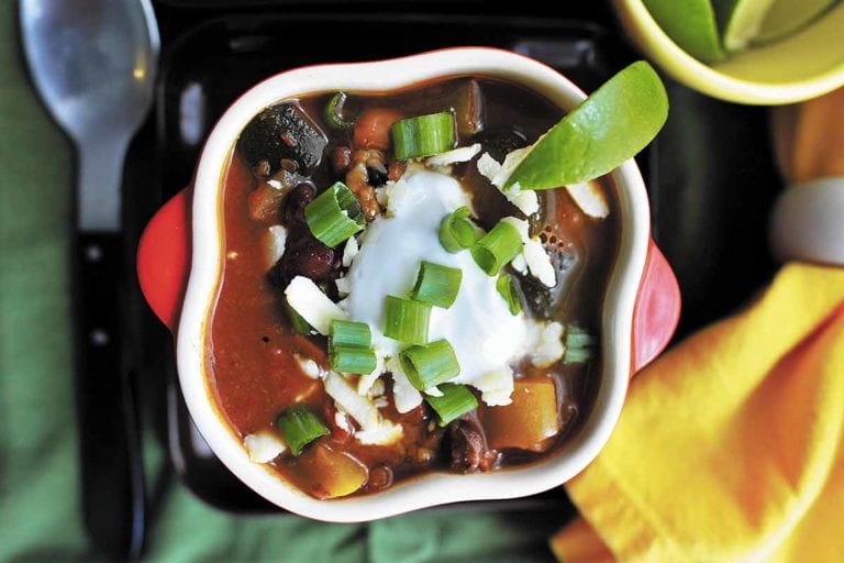 A white bowl with a slice of lime on the rim, filled with smoky chipotle vegetarian chili, sour cream, and topped with green onions.