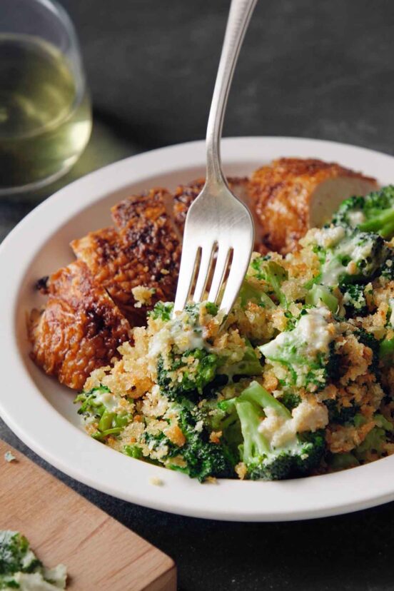 A close-up of the best broccoli casserole recipe served on a white plate next to sliced roast chicken, with a silver fork digging into the tender green broccoli, creamy blue cheese sauce, and crispy breadcrumb topping.