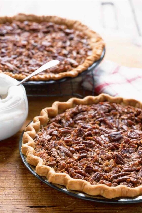 Two Southern pecan pies in pie plates with a bowl of whipped cream in between them.