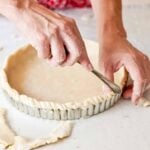Sweet pastry dough being placed in a fluted pie tin with hands fluting the edges.
