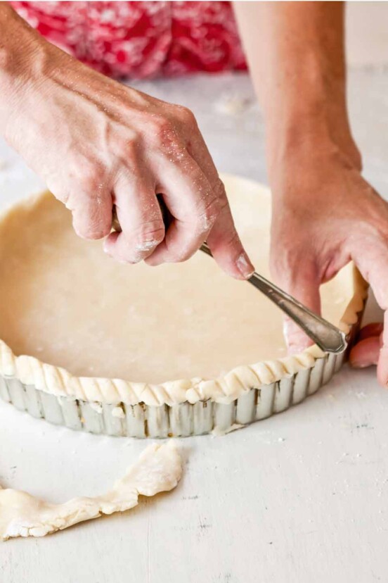 Sweet pastry dough being placed in a fluted pie tin with hands fluting the edges.
