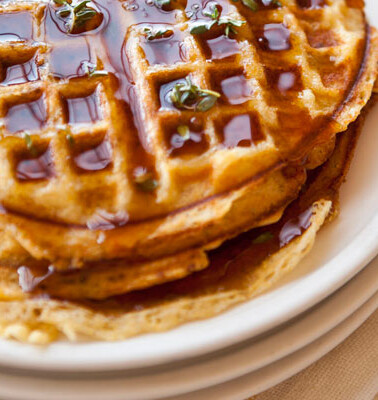 Cornmeal-bacon waffles with thyme infused maple syrup stacked on top of a pile of white plates.