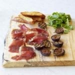 Slices of duck prosciutto, halved grilled figs, arugula, and a few crostini on a wooden serving board.