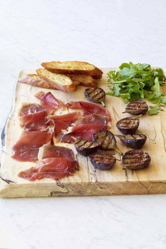 Slices of duck prosciutto, halved grilled figs, arugula, and a few crostini on a wooden serving board.