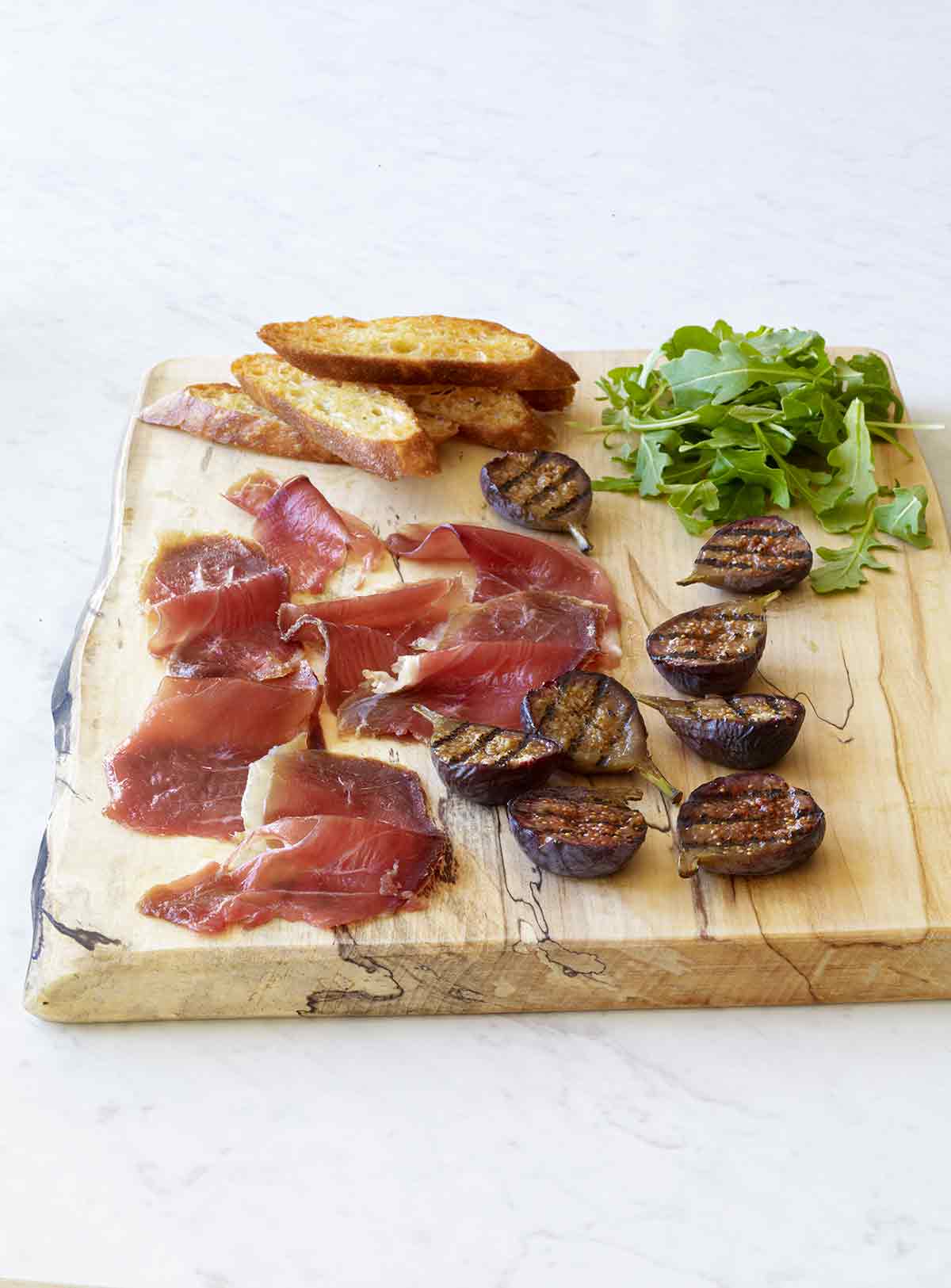 Slices of duck prosciutto, halved grilled figs, arugula, and a few crostini on a wooden serving board.