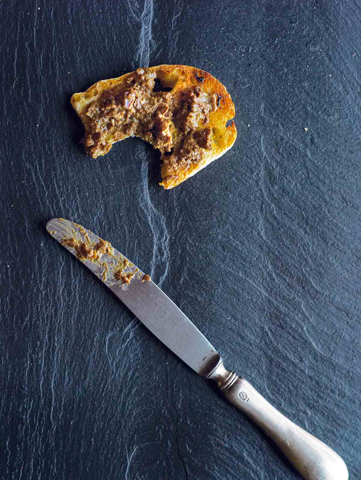 Sautéed chicken liver on a piece of toast with a bite out of it, on a piece of slate with a knife next to it. 