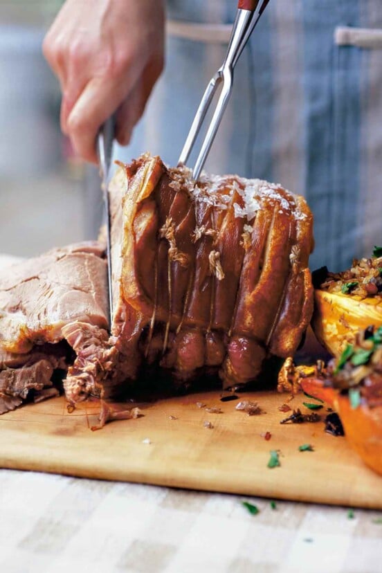 Slow-roasted pork shoulder on a cutting board being sliced by a person, flanked by two stuffed squash halves.