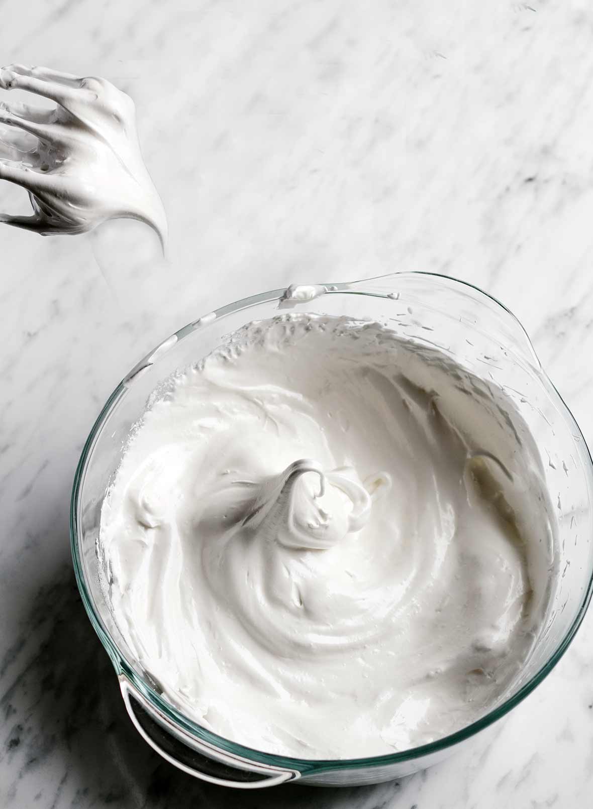Bowl of angel white icing in firm peaks on white marble, above is a whisk