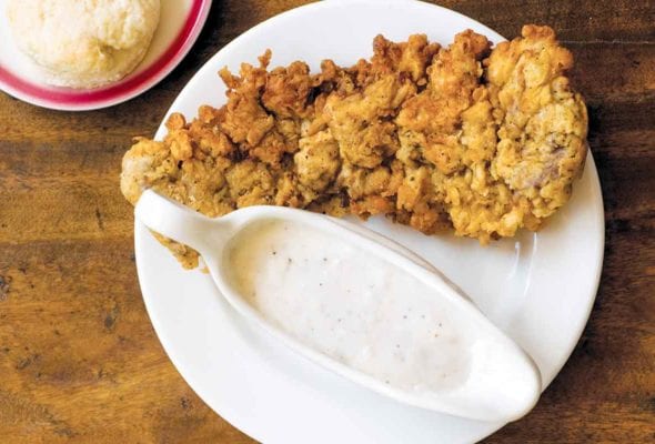 A piece of chicken fried steak with a dish of cream gravy beside it all on a white plate.